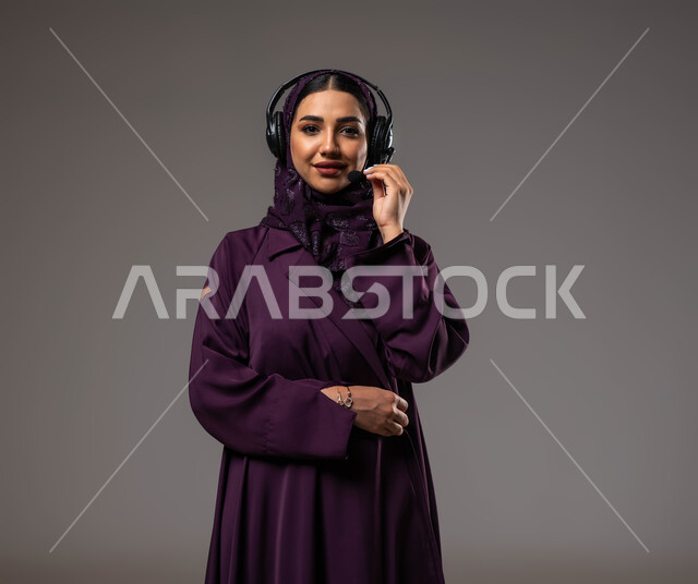 التواصل مع الزبائن لحل المشكلات والشكاوي، الإجابة والرد على الاستفسارات والاستعلامات, استخدام التقنيات المتطورة في العمل, بورتريه مقرب لامرأة عربية خليجية سعودية مبتسمة محجبة ترتدي العباءة تضع سماعات الرأس تعمل في قسم خدمة العملاء، خلفية رمادية