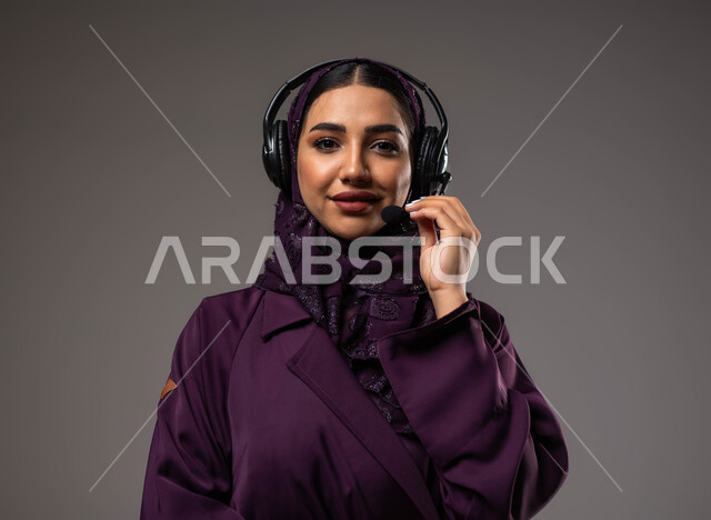 الإجابة والرد على الاستفسارات والاستعلامات, التواصل مع الزبائن لحل المشكلات والشكاوي، استخدام التقنيات المتطورة في العمل, بورتريه مقرب لامرأة عربية خليجية سعودية مبتسمة محجبة ترتدي العباءة تضع سماعات الرأس تعمل في قسم خدمة العملاء، خلفية رمادية