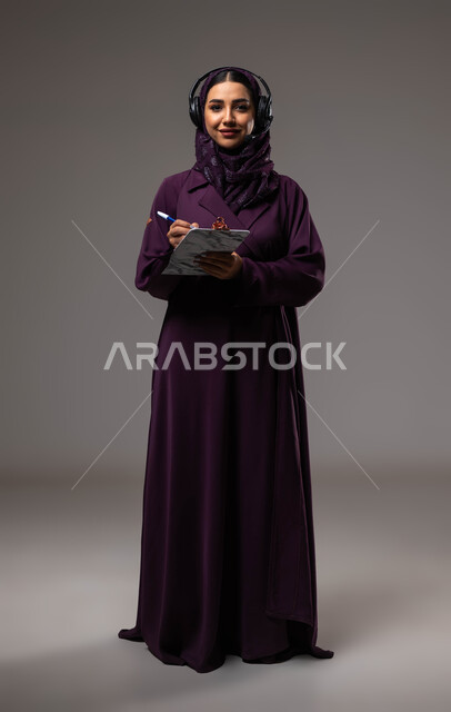 Taking and recording notes and complaints, following up and developing work, the concept of customer service, a portrait of a veiled Saudi Gulf Arab employee, smiling, wearing an abaya, wearing headphones on her head, looking at the camera, responding to inquiries and questions, full body photo, gray background