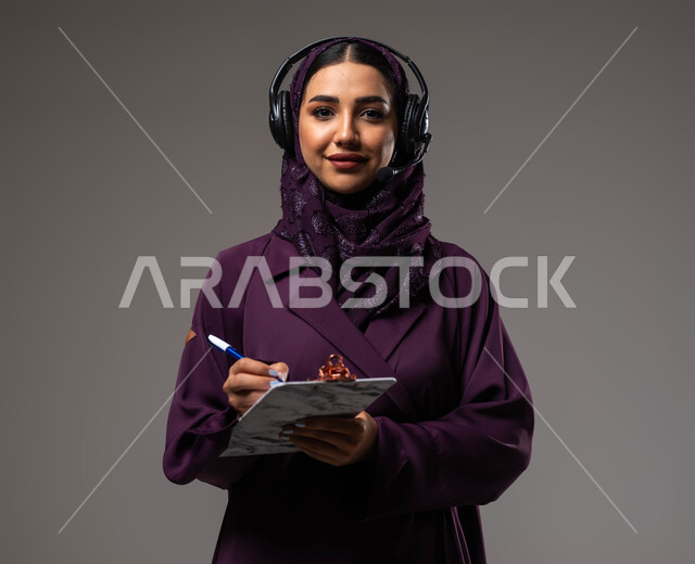 مفهوم خدمة العملاء، تدوين وتسجيل الملاحظات والشكاوي، متابعة وتطوير العمل، بورتريه مقرب لموظفة عربية خليجية سعودية محجبة مبتسمة ترتدي العباءة تضع سماعات الهيدفون على رأسها تنظر إلى الكاميرا، الرد على الاستفسارت والتساؤلات، خلفية رمادية