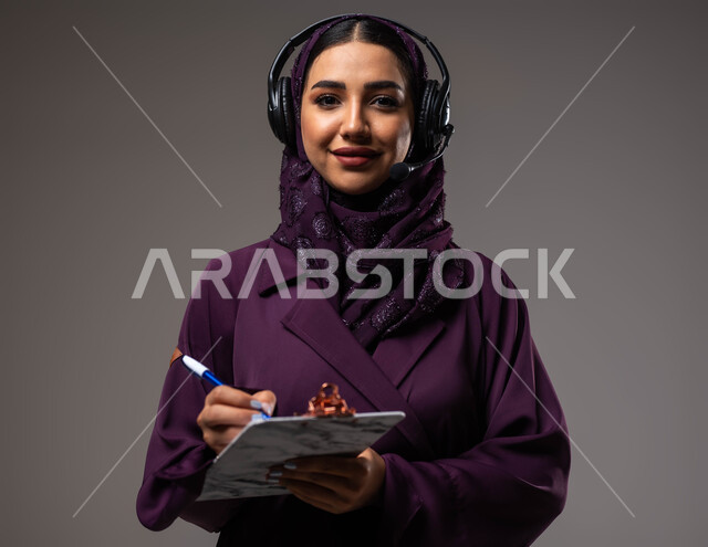 مفهوم خدمة العملاء، تدوين وتسجيل الملاحظات والشكاوي، متابعة وتطوير العمل، بورتريه مقرب لموظفة عربية خليجية سعودية محجبة مبتسمة ترتدي العباءة تضع سماعات الهيدفون على رأسها تنظر إلى الكاميرا، الرد على الاستفسارت والتساؤلات، خلفية رمادية