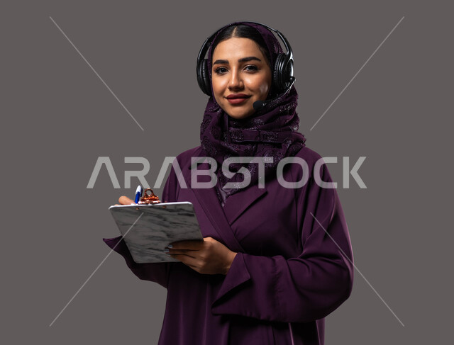 متابعة وتطوير العمل، مفهوم خدمة العملاء، تدوين وتسجيل الملاحظات والشكاوي، بورتريه مقرب لموظفة عربية خليجية سعودية محجبة مبتسمة ترتدي العباءة تضع سماعات الهيدفون على رأسها تقوم بالكتابة على الاوراق، الرد على الاستفسارت والتساؤولات، خلفية رمادية