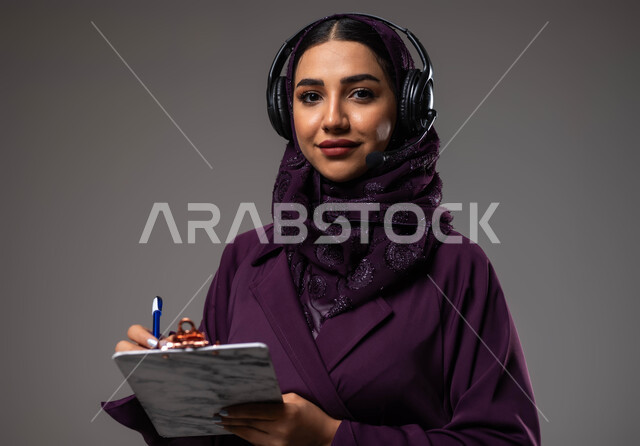 متابعة وتطوير العمل، مفهوم خدمة العملاء، تدوين وتسجيل الملاحظات والشكاوي، بورتريه مقرب لموظفة عربية خليجية سعودية محجبة مبتسمة ترتدي العباءة تضع سماعات الهيدفون على رأسها تقوم بالكتابة على الاوراق، الرد على الاستفسارت والتساؤولات، خلفية رمادية