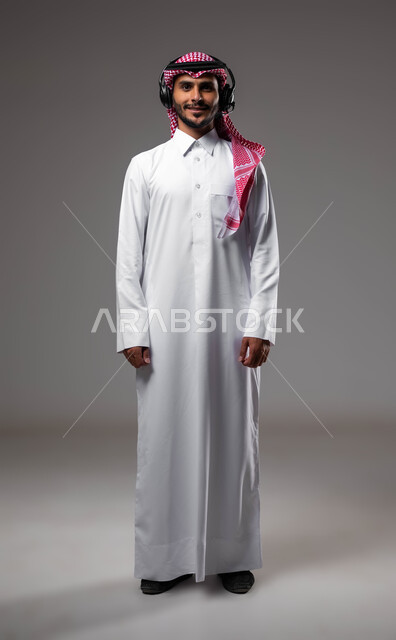 دمج التقنيات المتطورة مع العمل, النظر الى الكاميرا بتعابير الثقة بالنفس، الرد على الاستفسارات والاسئلة, بورتريه لشاب عربي خليجي سعودي يرتدي الزي التقليدي يضع الهيدفون يعمل في قسم خدمة العملاء، التواصل مع الزبائن لحل المشكلات، صورة الجسم بالكامل، خلفية رمادية