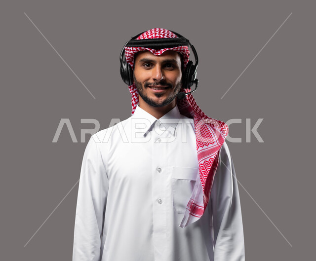 النظر الى الكاميرا بتعابير السرور والثقة بالنفس، الاجابة على الاستفسارات والاستعلامات, بورتريه مقرب لشاب عربي خليجي سعودي يرتدي الزي التقليدي يضع الهيدفون يعمل في قسم خدمة العملاء، دمج التقنيات المتطورة مع العمل, التواصل مع الزبائن لحل المشكلات، خلفية رمادية