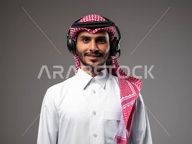 النظر الى الكاميرا بتعابير السرور والثقة بالنفس، الاجابة على الاستفسارات والاستعلامات, بورتريه مقرب لشاب عربي خليجي سعودي يرتدي الزي التقليدي يضع الهيدفون يعمل في قسم خدمة العملاء، دمج التقنيات المتطورة مع العمل, التواصل مع الزبائن لحل المشكلات، خلفية رمادية