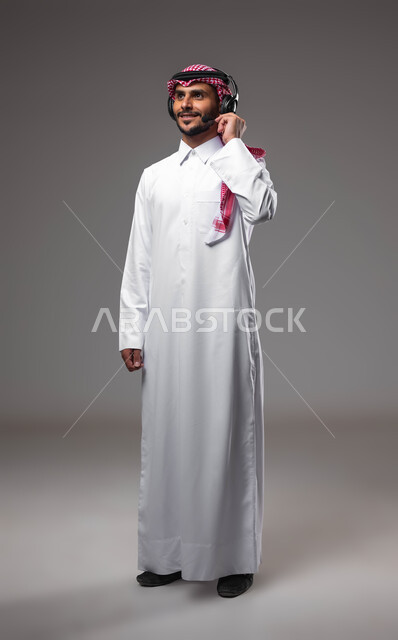استخدام سماعات الرأس والتقنيات المتطورة في العمل, الرد على الاستفسارات والاستعلامات, بورتريه لرجل عربي خليجي سعودي مبتسم يرتدي الشماغ والثوب التقليدي يضع الهيدفون يعمل في قسم خدمة العملاء، التواصل مع الزبائن لحل المشكلات والشكاوي، صورة الجسم بالكامل، خلفية رمادية