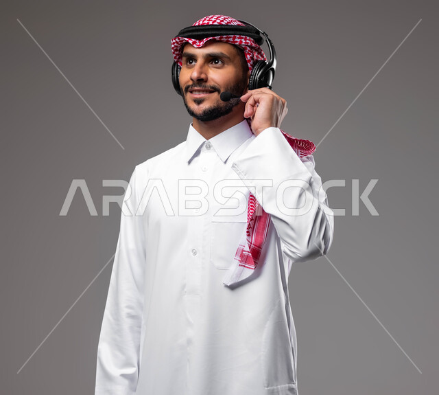 الرد والاجابة على الاستفسارات والاستعلامات, بورتريه مقرب لشاب عربي خليجي سعودي مبتسم يرتدي الشماغ والثوب التقليدي يضع الهيدفون يعمل في قسم خدمة العملاء، استخدام سماعات الرأس والتقنيات المتطورة في العمل, التواصل مع الزبائن لحل المشكلات والشكاوي، خلفية رمادية