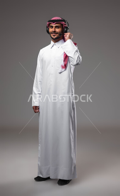 الرد على الاستفسارات والاستعلامات, استخدام سماعات الرأس والتقنيات المتطورة في العمل, بورتريه لرجل عربي خليجي سعودي مبتسم يرتدي الشماغ والثوب التقليدي يضع الهيدفون يعمل في قسم خدمة العملاء، التواصل مع الزبائن لحل المشكلات والشكاوي، صورة الجسم بالكامل، خلفية رمادية