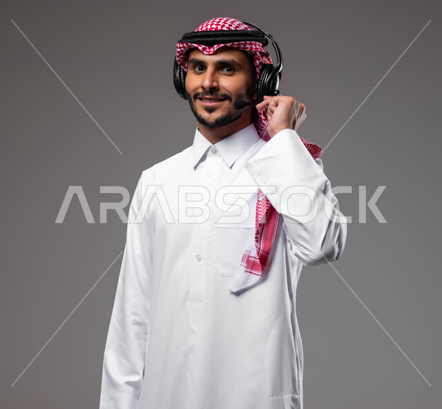 الإجابة والرد على الاستفسارات والاستعلامات, استخدام سماعات الرأس والتقنيات المتطورة في العمل, بورتريه مقرب لرجل عربي خليجي سعودي مبتسم يرتدي الشماغ والثوب التقليدي يضع الهيدفون يعمل في قسم خدمة العملاء، التواصل مع الزبائن لحل المشكلات والشكاوي، خلفية رمادية