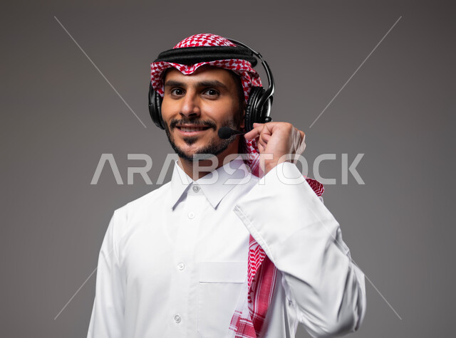 Communicating with customers to solve problems and complaints, answering and responding to inquiries and inquiries, using headphones and advanced technologies at work, a close-up portrait of a smiling Saudi Arabian Gulf man wearing a shemagh and traditional thobe with a headphone on, working in the customer service department, gray background
