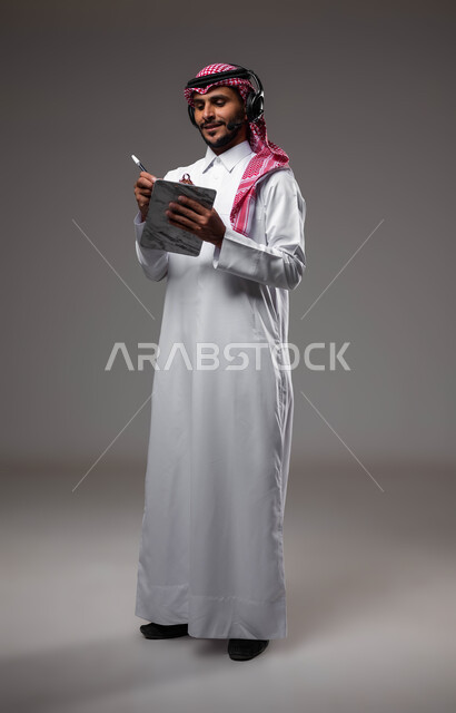 العمل في قسم خدمة العملاء، تدوين وتسجيل الملاحظات والشكاوي، بورتريه مقرب لموظف عربي خليجي سعودي مبتسم يرتدي الشماغ والثوب التقليدي يضع سماعات الهيدفون على رأسه يقوم بالكتابة على الاوراق، الرد على الاستفسارت والتساؤولات، صورة لكامل الجسم، خلفية رمادية