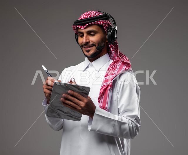 متابعة وتطوير العمل، تدوين وتسجيل الملاحظات والشكاوي، بورتريه مقرب لموظف عربي خليجي سعودي مبتسم يرتدي الشماغ والثوب التقليدي يضع سماعات الهيدفون على رأسه يقوم بالكتابة على الاوراق، الرد على الاستفسارت والتساؤولات، مفهوم خدمة العملاء، خلفية رمادية