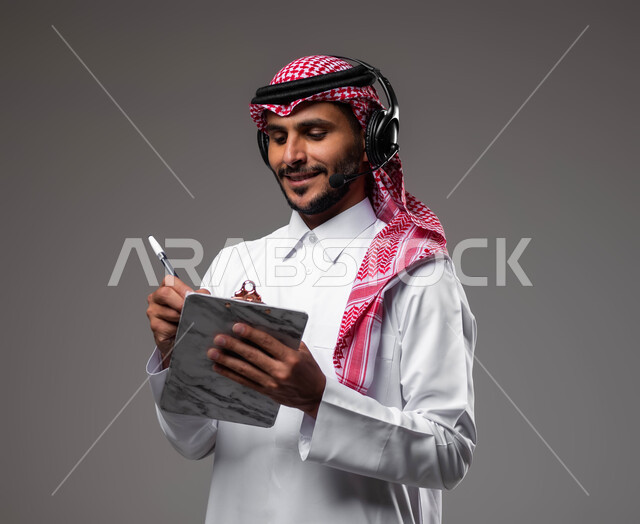 تدوين وتسجيل الملاحظات والشكاوي، بورتريه مقرب لموظف عربي خليجي سعودي مبتسم يرتدي الشماغ والثوب التقليدي يضع سماعات الهيدفون على رأسه يقوم بالكتابة على الاوراق، الرد على الاستفسارت والتساؤولات، متابعة وتطوير العمل، مفهوم خدمة العملاء، خلفية رمادية