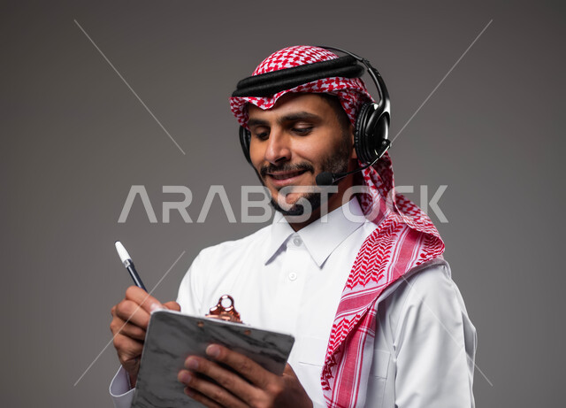 تسجيل وتدوين الملاحظات والشكاوي، بورتريه مقرب لموظف عربي خليجي سعودي مبتسم يرتدي الشماغ والثوب التقليدي يضع سماعات الهيدفون على رأسه يقوم بالكتابة على الاوراق، الرد على الاستفسارت والتساؤولات، متابعة وتطوير العمل، مفهوم خدمة العملاء، خلفية رمادية