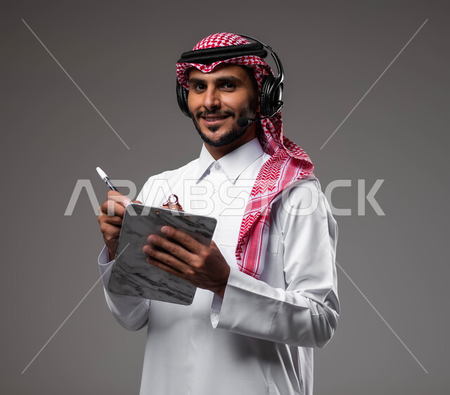 مفهوم خدمة العملاء، متابعة وتطوير العمل، تدوين وتسجيل الملاحظات والشكاوي، بورتريه مقرب لموظف عربي خليجي سعودي مبتسم يرتدي الشماغ والثوب التقليدي يضع سماعات الهيدفون على رأسه يقوم بالكتابة على الاوراق، الرد على الاستفسارت والتساؤولات، خلفية رمادية