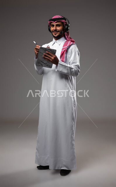 العمل في قسم خدمة العملاء، تدوين وتسجيل الملاحظات والشكاوي، بورتريه مقرب لموظف عربي خليجي سعودي مبتسم يرتدي الشماغ والثوب التقليدي يضع سماعات الهيدفون على رأسه يقوم بالكتابة على الاوراق، الرد على الاستفسارت والتساؤولات، صورة لكامل الجسم، خلفية رمادية