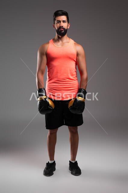 الاستعداد لبدء جولة المباراة، بورتريه لملاكم عربي خليجي سعودي يرتدي قفازات الملاكمة، الوقوف باستقامة بتعابير الثقة بالنفس، ممارسة رياضة الدفاع عن النفس، الملاكمة والمصارعة، نشاط بدني للحفاظ على الصحة واللياقة الجسدية، صورة الجسم بالطول الكامل، خلفية رمادية