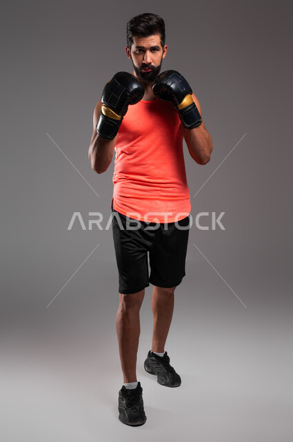 الملاكمة والمصارعة، التدرب على الحركات القتالية، بورتريه لملاكم عربي خليجي سعودي يرتدي قفازات الملاكمة  يمارس رياضة الدفاع عن النفس، نشاط بدني للحفاظ على الصحة واللياقة الجسدية، صورة الجسم بالطول الكامل، خلفية رمادية