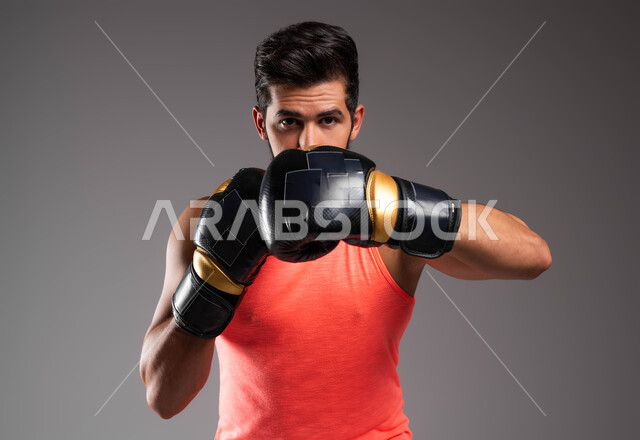 ممارسة رياضة الدفاع عن النفس، نشاط بدني للحفاظ على الصحة واللياقة الجسدية، بورتريه مقرب لملاكم عربي خليجي سعودي يرتدي قفازات الملاكمة يقوم بالتدرب على الحركات القتالية، المصارعة والملاكمة، حركات وتعابير الدفاع والهجوم، خلفية رمادية
