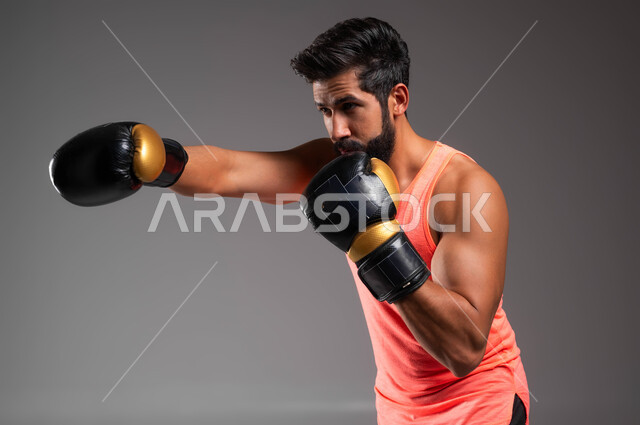 حركات وتعابير الدفاع والهجوم، ممارسة رياضة الدفاع عن النفس، نشاط بدني للحفاظ على الصحة واللياقة الجسدية، بورتريه مقرب لملاكم عربي خليجي سعودي يرتدي قفازات الملاكمة يقوم بالتدرب على الحركات القتالية، المصارعة والملاكمة، خلفية رمادية