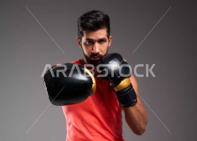 ممارسة رياضة الدفاع عن النفس، نشاط بدني للحفاظ على الصحة واللياقة الجسدية، بورتريه مقرب لملاكم عربي خليجي سعودي يرتدي قفازات الملاكمة يقوم بالتدرب على الحركات القتالية، المصارعة والملاكمة، حركات وتعابير الدفاع والهجوم، خلفية رمادية