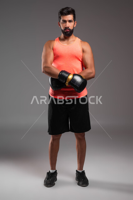 التدرب على حركات قتالية، بورتريه لملاكم عربي خليجي سعودي يرتدي قفازات الملاكمة يقف باستعداد واستقامة، ممارسة رياضة الدفاع عن النفس، الملاكمة والمصارعة، نشاط بدني للحفاظ على الصحة واللياقة الجسدية، صورة الجسم بالطول الكامل، خلفية رمادية