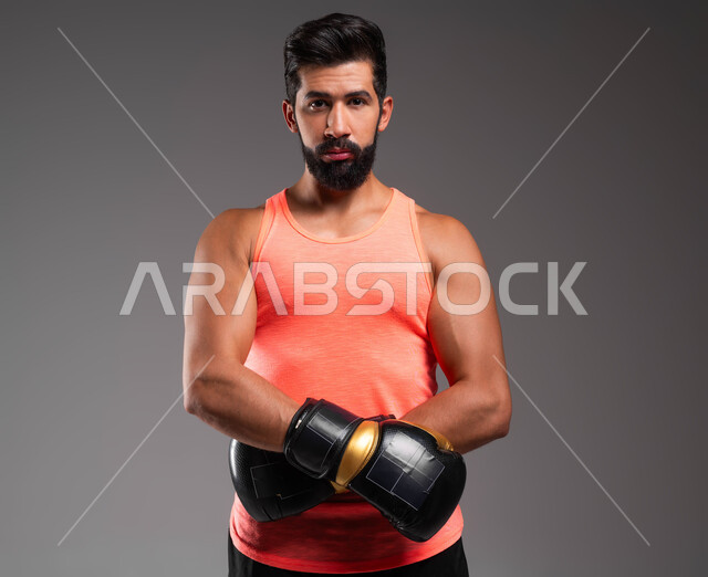 احتراف رياضة البوكسينغ، التدرب على فنون المصارعة، أنشطة بدنية للحفاظ على اللياقة الجسدية، بورتريه من الجانب لملاكم عربي خليجي سعودي يرتدي الملابس الرياضية وقفازات الملاكمة ينظر إلى الكاميرا بإيماءات الجدية، خلفية رمادية .