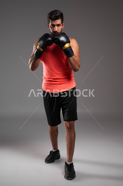 ممارسة رياضة البوكسينغ، نشاط بدني للحفاظ على الصحة واللياقة الجسدية، التدرب على فنون المصارعة، بورتريه لملاكم عربي خليجي سعودي يرتدي الملابس الرياضية وقفازات الملاكمة ينظر إلى الكاميرا بإيماءات الجدية، صورة الجسم بالطول الكامل، خلفية رمادية .