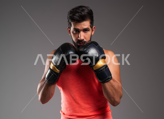 ممارسة رياضة البوكسينغ, التدرب على فنون المصارعة، بورتريه لملاكم عربي خليجي سعودي يرتدي الملابس الرياضية وقفازات الملاكمة ينظر إلى الكاميرا يرفع يديه أمام وجهه، نشاط بدني للحفاظ على الصحة واللياقة الجسدية، خلفية رمادية .