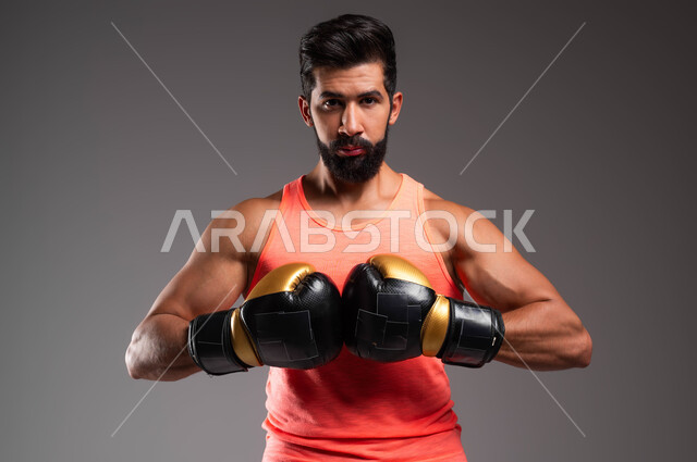 احتراف الحركات القتالية، نشاط بدني للحفاظ على الصحة واللياقة الجسدية، بورتريه لملاكم عربي خليجي سعودي يرتدي الملابس الرياضية وقفازات الملاكمة ينظر إلى الكاميرا بإيماءات الجدية، التدرب على فنون المصارعة, ممارسة رياضة البوكسينغ، خلفية رمادية .