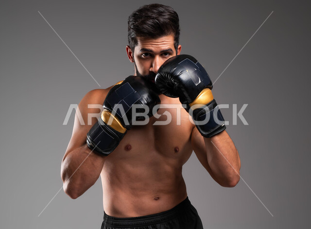 حركات التصدي وحماية الوجه، مفهوم الصحة واللياقة الجسدية، بورتريه لملاكم عربي خليجي سعودي يرتدي الشورت الرياضي وقفازات الملاكمة يقوم بالتدرب على فنون المصارعة، ممارسة رياضة البوكسينغ، خلفية رمادية