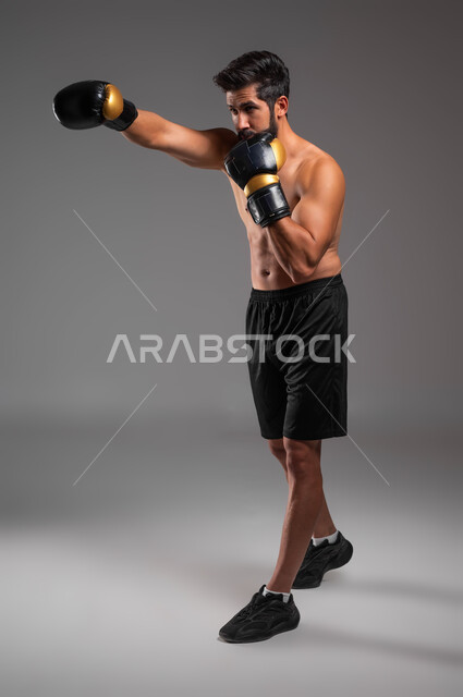 ممارسة رياضة البوكسينغ، حركات الهجوم والقتال، مفهوم الصحة واللياقة الجسدية، بورتريه لملاكم عربي خليجي سعودي يرتدي الشورت الرياضي وقفازات الملاكمة يقوم بالتدرب على فنون المصارعة, رياضات وأنشطة بدنية، صورة الجسم بالطول الكامل, خلفية رمادية .