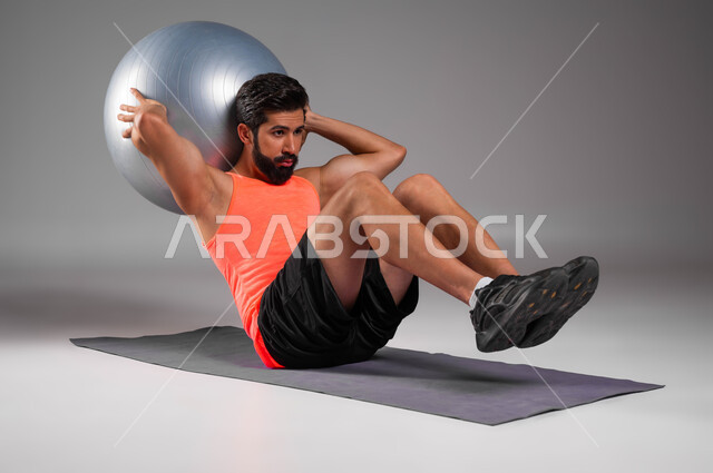 اللعب بكرة تمارين الايروبيك واللياقة البدنية، بورتريه للاعب عربي خليجي سعودي يرتدي الملابس الرياضية يقوم بممارسة التمارين الرياضية لشد عضلات البطن والمحافظة على الصحة الجسدية، مفهوم الصحة والرشاقه، أنشطة ورياضات شبابية، خلفية رمادية