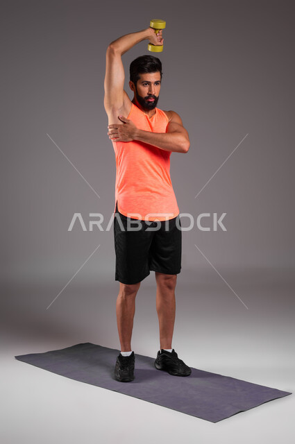 تمارين رفع الأثقال والأوزان الحديدية الثقيلة، بورتريه لشاب عربي خليجي سعودي يمارس التمارين الرياضية مع الدمبلز، نشاط بدني للمحافظة على الصحة واللياقة الجسدية، صورة الجسم بالكامل، كمال الأجسام وتقوية وبناء العضلات، أسلوب حياة صحي، خلفية رمادية