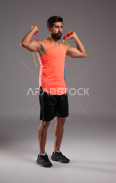 الاهتمام بالنشاط والحركة، أسلوب حياة صحي، نشاط بدني للمحافظة على الصحة واللياقة الجسدية، القيام بتمارين القفز على الحبل، بورتريه لشاب عربي خليجي سعودي يرتدي الملابس الرياضية يقوم بممارسة تمارين نط الحبل، صورة الجسم بالكامل، خلفية رمادية