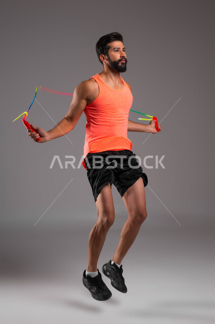 Physical activity to maintain health and physical fitness, doing jumping rope exercises, portrait of a Saudi Gulf Arab young man wearing sports clothing doing rope jumping exercises, interest in activity and movement, healthy lifestyle, full body image, gray background