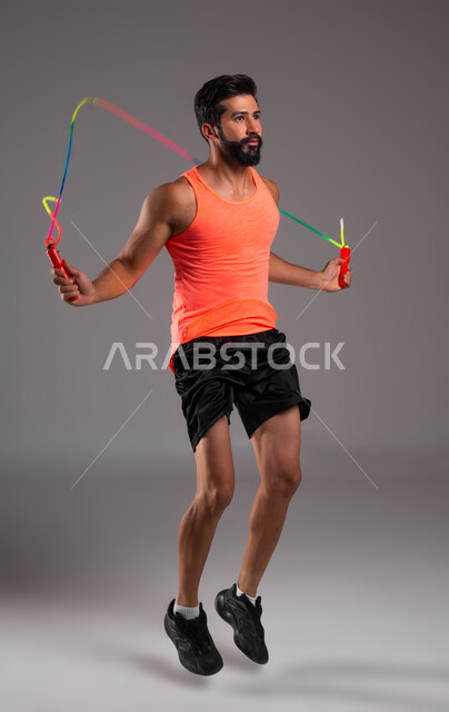 القيام بتمارين القفز على الحبل، بورتريه لشاب عربي خليجي سعودي يرتدي الملابس الرياضية يقوم بممارسة تمارين نط الحبل، نشاط بدني للمحافظة على الصحة واللياقة الجسدية، الاهتمام بالنشاط والحركة، أسلوب حياة صحي، صورة الجسم بالكامل، خلفية رمادية