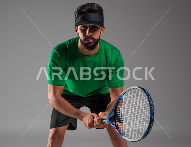 إيماءات تدل على الحماس والاندماج بالتمرين، رياضات شبابية وأنشطة ترفيهية، التدرب على إتقان لعبة البادل، إجراء تمرينات رياضة المضرب، بورتريه مقرب للاعب بادل عربي خليجي سعودي يرتدي ملابس رياضية يمسك المضرب بيديه، خلفية رمادية