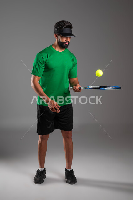 تعابير وإيماءات تدل على الحماس والاندماج بالتمرين، إجراء تمرينات رياضة المضرب، بورتريه للاعب بادل عربي خليجي سعودي يرتدي ملابس رياضية و يلعب بالكرة والمضرب، التدرب على إتقان لعبة البادل، أنشطة ترفيهية و رياضات شبابية، صورة الجسم بالكامل، خلفية رمادية
