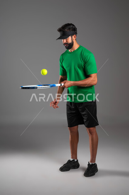 تعابير وإيماءات تدل على الحماس والاندماج بالتمرين، إجراء تمرينات رياضة المضرب، بورتريه للاعب بادل عربي خليجي سعودي يرتدي ملابس رياضية و يلعب بالكرة والمضرب، التدرب على إتقان لعبة البادل، أنشطة ترفيهية و رياضات شبابية، صورة الجسم بالكامل، خلفية رمادية