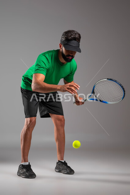Practicing fun sports and physical activities, a portrait of a Saudi Gulf Arab young man wearing sports clothes and holding a tennis racket in his hand, playing quickly and with concentration, maintaining sports and physical fitness, gray background