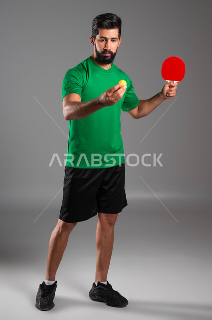 اللعب باتقان وتركيز، ممارسة رياضات وانشطة بدنية شبابية ممتعة، بورتريه للاعب عربي خليجي سعودي يرتدي ملابس رياضية يحمل بيده مضرب وكرة تنس الطاولة، المحافظة على الرياضة والرشاقة الجسدية، صورة الجسم بالطول الكامل، خلفية رمادية