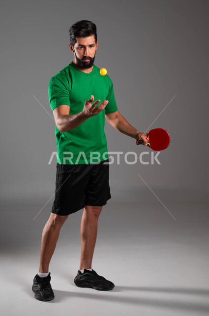 Focusing on playing and mastering it, practicing fun youth sports and physical activities, portrait of a Saudi Arabian Gulf player wearing sports clothing holding a racket and a table tennis ball in his hand, maintaining sports and physical fitness, full-length body image, gray background