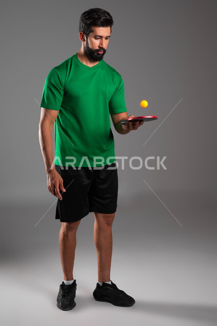 اللعب باتقان وتركيز، ممارسة رياضات وانشطة بدنية شبابية ممتعة، بورتريه للاعب عربي خليجي سعودي يرتدي ملابس رياضية يحمل بيده مضرب وكرة تنس الطاولة، المحافظة على الرياضة والرشاقة الجسدية، صورة الجسم بالطول الكامل، خلفية رمادية
