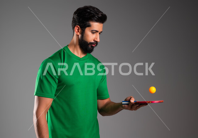 ممارسة رياضات وانشطة بدنية شبابية ممتعة، بورتريه مقرب للاعب عربي خليجي سعودي يرتدي ملابس رياضية يحمل بيده مضرب وكرة تنس الطاولة، اللعب باتقان وتركيز، المحافظة على الرياضة والرشاقة الجسدية، خلفية رمادية