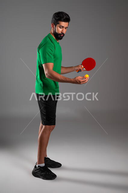 ممارسة رياضات وانشطة بدنية ممتعة، المحافظة على الرياضة والرشاقة الجسدية، اللعب بسرعة وتركيز، بورتريه لشاب عربي خليجي سعودي يرتدي ملابس رياضية يحمل بيده مضرب تنس الطاولة، صورة لكامل الجسم، خلفية رمادية