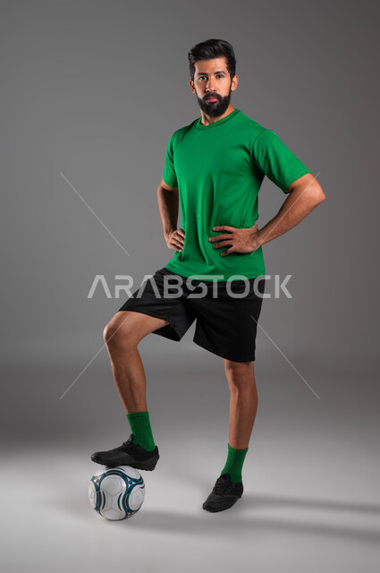 الوقوف باستقامة والنظر إلى الكاميرا، ممارسة هواية اللعب بكرة القدم، بورتريه للاعب عربي سعودي خليجي يرتدي الملابس الرياضية يقف باستعداد لبدء المباراة، احتراف الرياضات الجماعية، صورة الجسم بالطول الكامل، خلفية رمادية .
