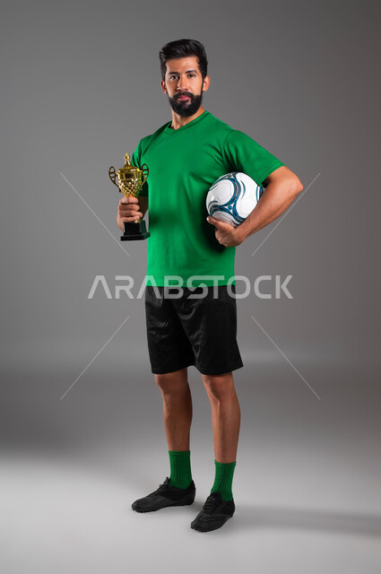 ممارسة الرياضات البدنية الممتعة، احتراف رياضة كرة القدم، النظر إلى الكاميرا بإيماءات الفوز والانتصار، الشعور بالفرح والسرور، بورتريه للاعب عربي سعودي خليجي يرتدي الملابس الرياضية يحمل بيده كأس البطولة والكرة، صورة لكامل الجسم، خلفية رمادية .