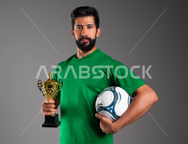 Practicing fun physical sports, mastering the sport of football, looking at the camera with gestures of victory and triumph, feeling joy and pleasure, a close-up portrait of a smiling Saudi Arabian Gulf player wearing sports clothes, holding a championship cup and ball in his hand, gray background.